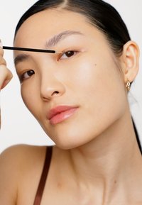 Modell som applicerar produkt med en tunn svart applikator. Har slät hy, naturlig makeup och små guldsmycken. Ljust bakgrund.