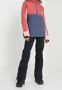 Kolorowa kurtka narciarska z różowymi i fioletowymi panelami, z kieszenią z przodu i kapturem wykończonym futrem. Dopasowana do czarnych spodni narciarskich i butów.