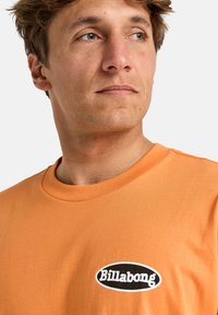 Oranžové bavlněné tričko s černým oválným logem s nápisem "Billabong" v bílé barvě. Klasický kulatý výstřih a krátké rukávy. Hladká textura.