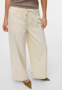 Krēmkrāsas platās bikses ar elastīgu jostu un auklu. Iespējas sānu kabatas un gluda auduma tekstūra. Kombinētas ar smailiem apaviem.