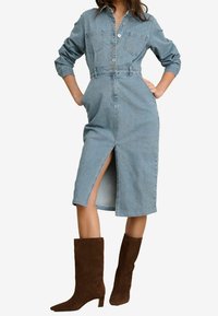 Abito camicia in denim blu chiaro con maniche lunghe, chiusura a bottoni sul davanti, due tasche sul petto e uno spacco frontale. Abbinato a stivali al ginocchio in suede marrone.