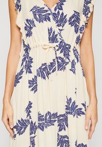 Robe crème avec motif de feuilles bleues, col en V, manches courtes à volants et lien à nouer à la taille, portée par une personne les bras détendus le long du corps.