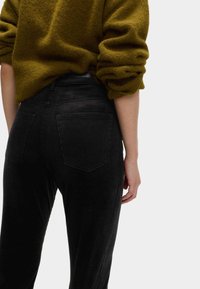 Schwarze Cordhosen mit hoher Taille, ausgestattet mit zwei Gesäßtaschen und einer strukturierten Oberfläche. Kombiniert mit einem lockeren, olivgrünen Pullover.