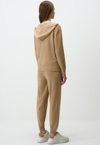 Tanskleurige pullover hoodie met een ontspannen pasvorm, geribbelde manchetten en tailleband, gecombineerd met bijpassende taps toelopende broeken en neutrale sneakers.