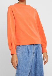 Oranges Sweatshirt mit rundem Halsausschnitt und langen, leicht puffigen Ärmeln. Kombiniert mit lockeren blauen Jeanshosen.