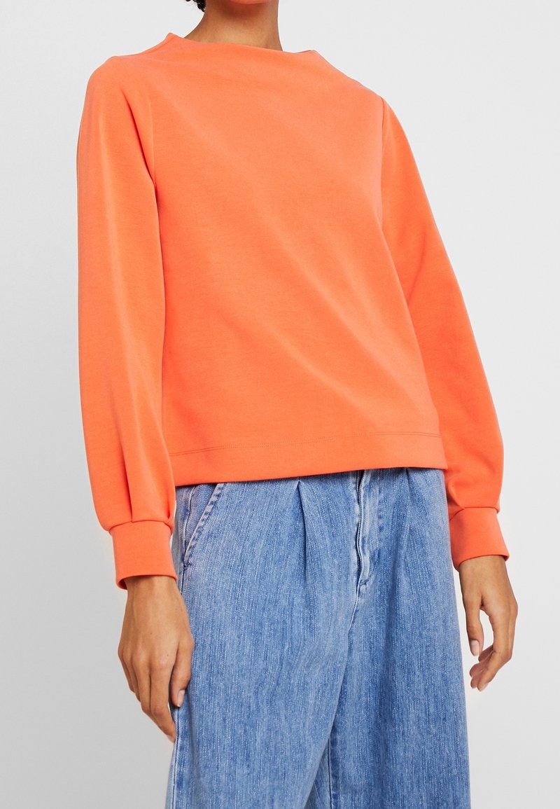 Oranges Sweatshirt mit rundem Halsausschnitt und langen, leicht puffigen Ärmeln. Kombiniert mit lockeren blauen Jeanshosen.