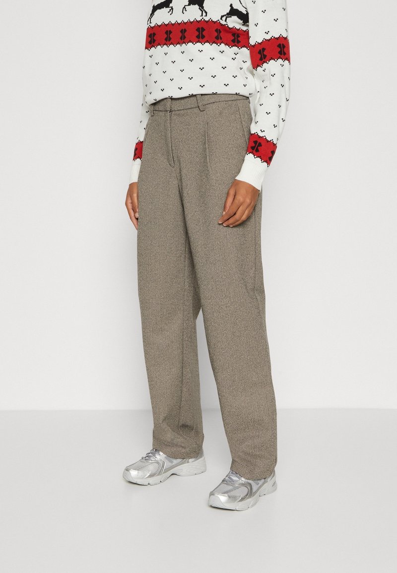 Personne portant un pantalon beige à jambes larges, des baskets argentées et un pull blanc avec des motifs rouges et noirs, debout devant un fond blanc.