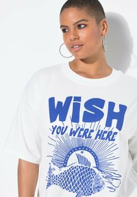 Witte oversized t-shirt met blauwe grafische tekst "WISH YOU WERE HERE", vissen en zonnestraalontwerp. Zachte katoenen textuur.
