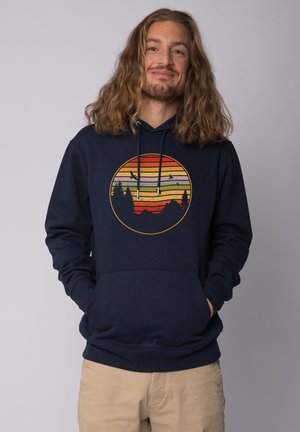 Navy blue hoodie with a circular graphic of layered stripes in red, orange, green, and yellow, depicting mountains and trees. Cotton blend fabric.