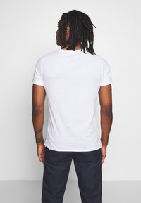 Hvid bomulds T-shirt med korte ærmer, rund halsudskæring, lidt tætsiddende form, båret sammen med marineblå bukser, synlig tatovering på armen.