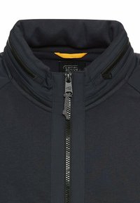 Donkergrijze vest met ritssluiting en hoge kraag, met een contrasterende gele binnenkraag en een textuurdetail aan de ritshanger. Gladde stofstructuur.