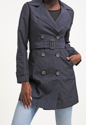 Manteau trench à double boutonnage bleu marine foncé avec ceinture, larges revers et poignets boutonnés, porté sur un haut noir et un jean bleu clair.