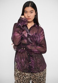 Blusa viola traslucida con motivo a foglia, dettagli arricciati lungo il davanti con bottoni, e bottoni dorati. Abbinata a pantaloni con stampa leopardata.