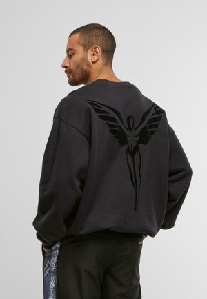 Man wearing black sweatshirt with large black winged figure design on back, facing sideways with hands in pants pockets.