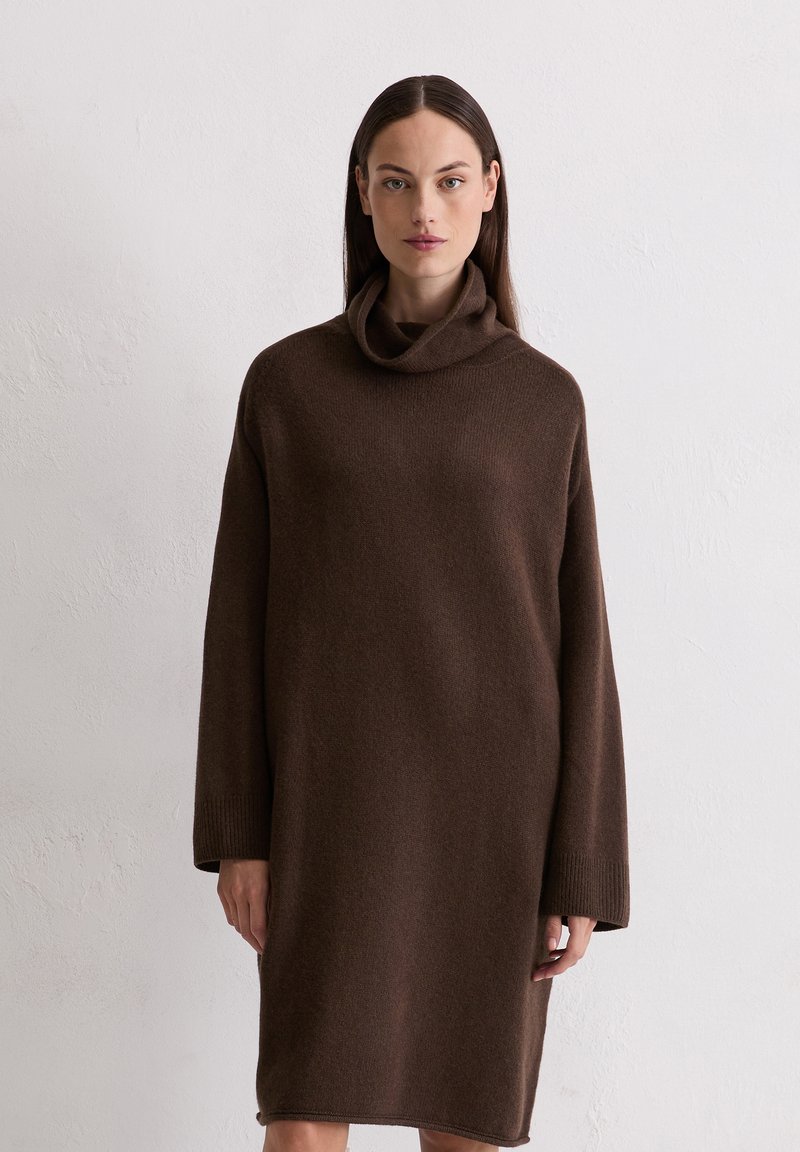 Frau mit langen, dunklen Haaren, die ein lockeres, langärmliges, braunes Rollkragenkleid trägt und vor einer schlichten hellen Wand steht.