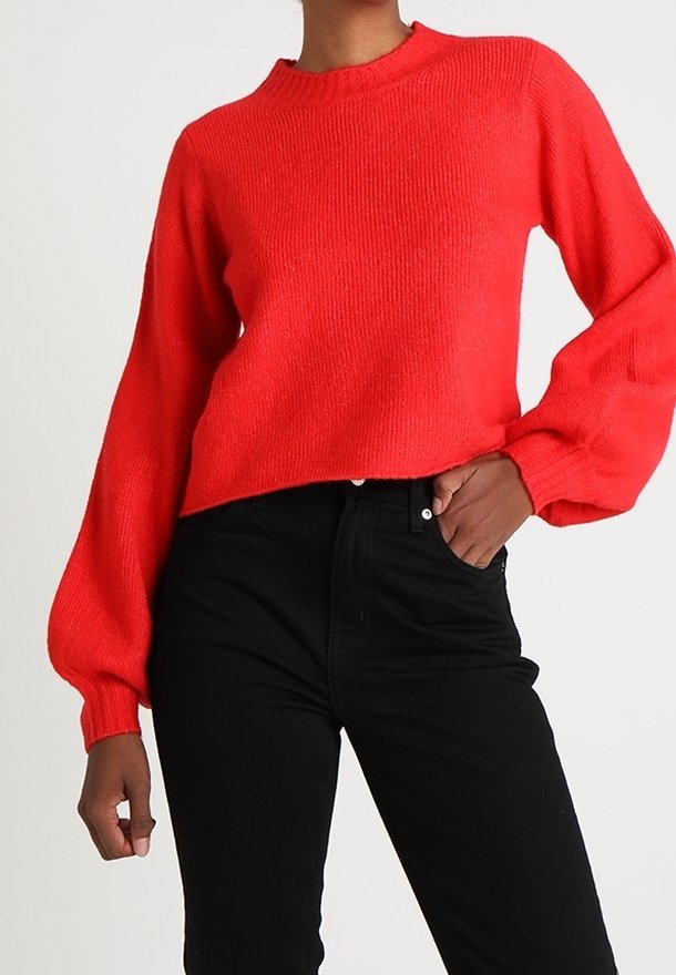 Punainen neulottu neule, jossa pitkät, liehulahkeiset hihat ja pyöreä pääntie, yhdistetty mustiin korkeavyötäröisiin farkkuihin. Pehmeä tekstuuri ja rento istuvuus.