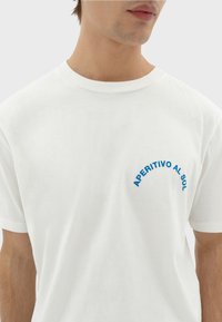 Weißes Baumwoll-T-Shirt mit einem blauen Grafikdruck, der den Schriftzug "APERITIVO AL SOL" zeigt und in einer geschwungenen Anordnung auf der Brust platziert ist.