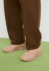 Lys rosa sneakers med teksturert semsket skinn og mesh-overdel, rund tå, snøring, og robuste gummisåler. Båret med brune baggy bukser.