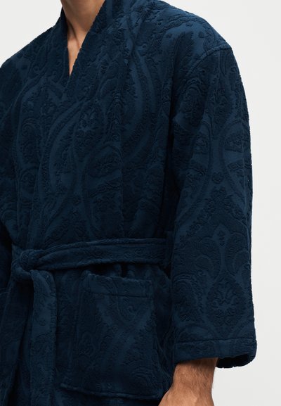 Homme portant un peignoir texturé bleu foncé avec une ceinture nouée à la taille et une poche à l'avant.