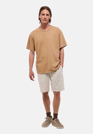 Jeune homme debout portant un t-shirt marron ample, un short beige, des chaussettes marron et des baskets beiges sur un fond blanc uni.