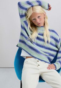 Suéter a rayas en verde menta, azul y morado de lana con puños acanalados, combinado con pantalones blancos de cintura alta y una silla turquesa.