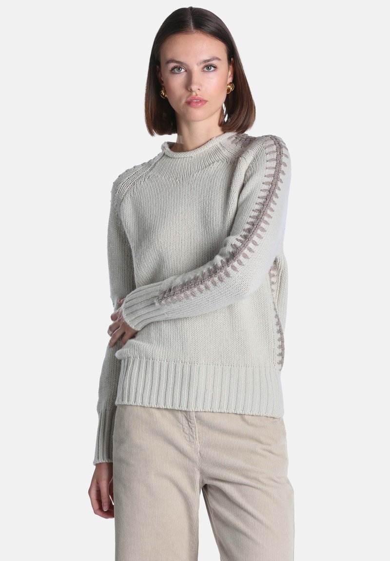 Jeune femme aux cheveux courts et foncés porte un pull en maille gris clair avec un détail en relief sur les manches et un pantalon en velours côtelé beige.