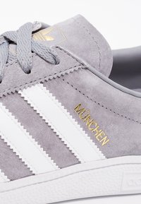 Szara zamszowa tenisówka z białymi skórzanymi paskami i złotym logo "MÜNCHEN". Posiada teksturowaną cholewkę, okrągłe sznurówki oraz gumową podeszwę.