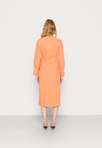 Vestido laranja, até ao joelho, com mangas longas, apresentando um fecho com zíper nas costas e tecido texturizado. Par de sandálias de dedo aberto com salto baixo.