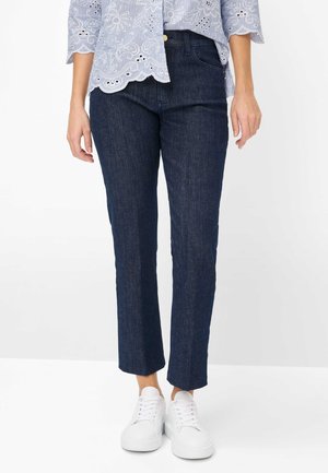 Kvinde iført mørkeblå jeans med lige ben, en lyseblå broderet bluse og hvide sneakers, stående på en hvid baggrund.