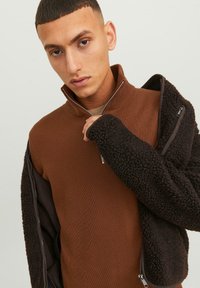 Hombre joven con el cabello corto, vistiendo un suéter marrón de canalé con cremallera y una chaqueta texturizada marrón oscuro colgada de un hombro.
