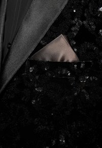 Zwarte paillettenjas met een satijnen revers, voorzien van een lichtbruine, gemêleerde zakdoek die in het borstzakje is gestoken.