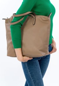 Beige leather tote bag with a textured finish, top zipper, and dual handles, held against a green sweater. Features black edging.