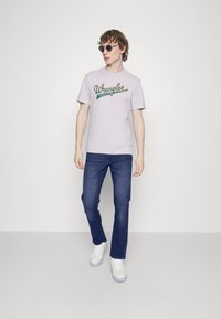 Svijetlo lavanda majica s "Wrangler" logom, tamnoplave traperice i bijeli tenisice. Model nosi okrugle crne sunčane naočale. Jednostavan, ležeran izgled.