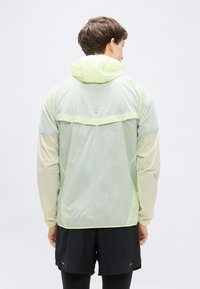 Ελαφρύ, διαφανές αδιάβροχο σε ανοιχτό πράσινο, με κουκούλα, πίσω τσέπη με φερμουάρ και αντανακλαστικές λεπτομέρειες. Μακριά μανίκια με ελαστικές μανσέτες.