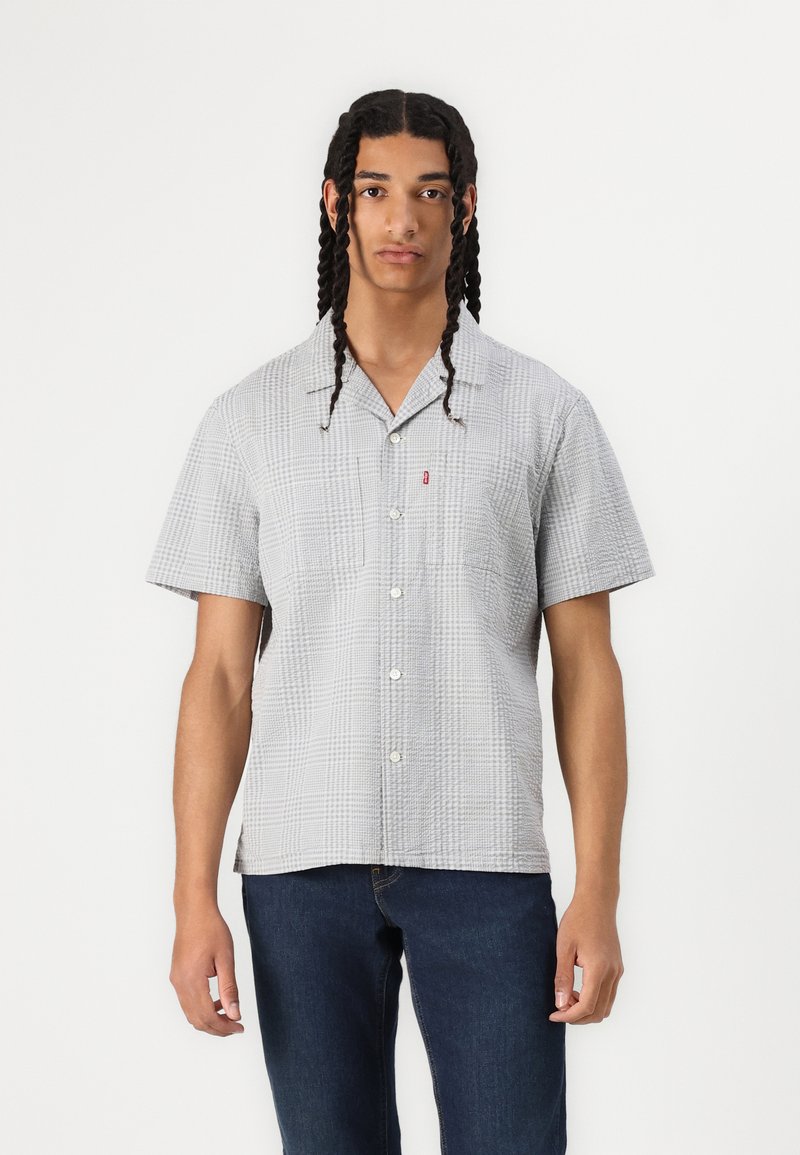 Jeune homme avec de longs cheveux tressés portant une chemise à carreaux à manches courtes gris clair et un jean bleu foncé, se tenant devant un fond blanc.