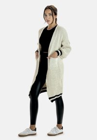 Stickad kofta i creme med svarta detaljer, öppen fram, långa ärmar och framfickor, ihopparad med svarta leggings och vita sneakers.