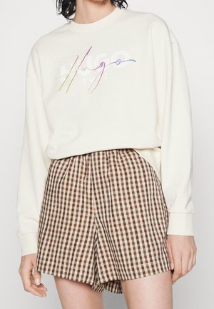 Henkilö, jolla on valkoinen pitkähihaisen paita, jossa värikäs käsinkirjoitettu Hugo-logo, ja korkeavyötäröiset, ruskeat ja beiget ruudulliset shortsit.