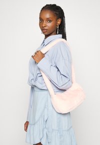 Bolsa de ombro de pelúcia rosa, com formato arredondado, textura suave e uma alça longa, usada sobre uma camisa azul clara e uma saia de ganga.