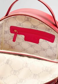 Interno di uno zaino in pelle rossa che presenta una tasca con zip, foderato in beige chiaro con un motivo a logo ripetuto, e dettagli della zip in tono oro.