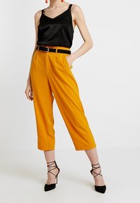 Černá saténová košilka, vysoce pasové žluté zkrácené kalhoty s plisováním, černý opasek a černé lodičky na jehlovém podpatku s ankletovými stuhami.