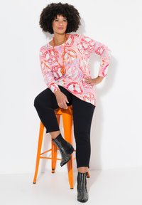 Langärmliges Oberteil mit einem rosa Friedenzeichen-Muster, kombiniert mit schwarzen Leggings und Nieten-Stiefeletten, die auf einem orangefarbenen Barhocker sitzen.