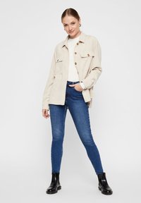 Beige Cordjacke mit zwei vorderen Taschen, braunen Knöpfen, kombiniert mit engen blauen Jeans und schwarzen Stiefeletten. Glatte Textur, lässiges Design.