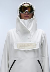 White waterproof jacket with a high collar, adjustable hood, front logo patch, and black zipper pockets. Model wearing ski goggles.