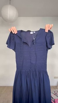 Marineblå kjole med korte pufærmer, rynket overdel, samlet nederdel, holdt op med hænder i et lyst rum med en loftslampe ovenover.