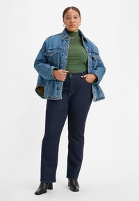 Jaqueta de ganga com lavagem azul, corte oversized; suéter de gola alta às riscas verdes e pretas; jeans de cintura alta azul-escuro; botas pretas de salto bloco.