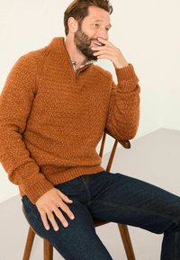 Rustfarbenes Strickpullover mit einem halben Reißverschluss-Kragen, strukturiertem Muster und gerippten Bündchen. Kombiniert mit dunklen Jeans sitzt er auf einem Holzstuhl.