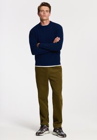 Námořnický pletený svetr, olivové žebrované kalhoty a vzorované tenisky. Model stojí se zkříženýma rukama na jednobarevném pozadí.