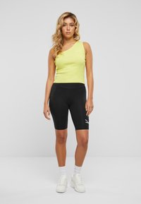 Limegrünes, geripptes Tanktop mit einem Schulterdesign, kombiniert mit hoch taillierten schwarzen Radlerhosen mit einem weißen Logodetail. Weiße Sneakers.