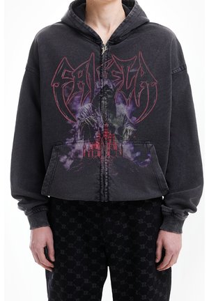 Osoba ubrana w ciemną bluzę na zamek z grafiką postaci w kapturze nad czerwonym zamkiem oraz stylizowanym napisem, w połączeniu z ciemnymi spodniami z wzorem.