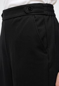 Gros plan sur un pantalon noir avec une poche latérale et une taille haute dotée d'un rabat à bouton réglable, porté avec une chemise blanche.